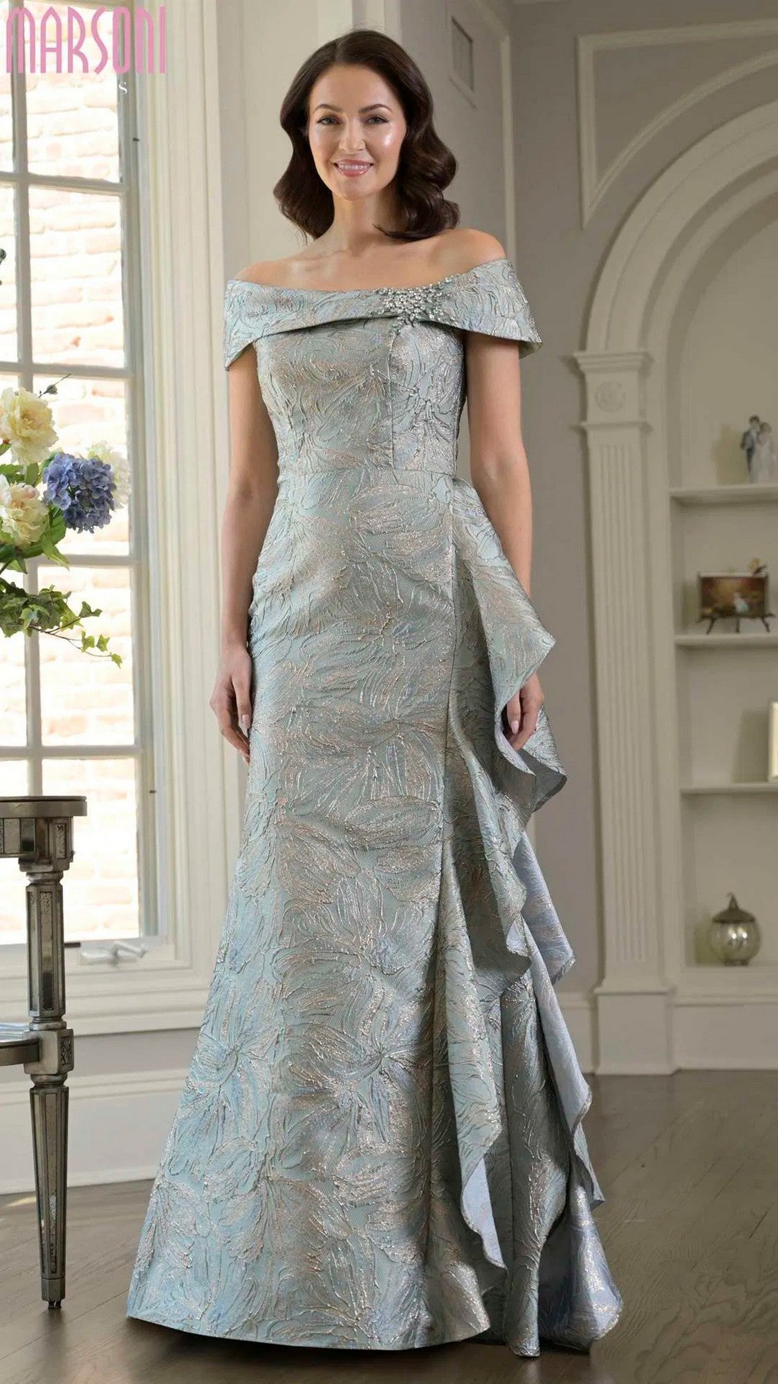 A woman in a sage Marsoni MV1381 long formal dress with an elegant off-the-shoulder neckline stands indoors near a window and floral arrangement.